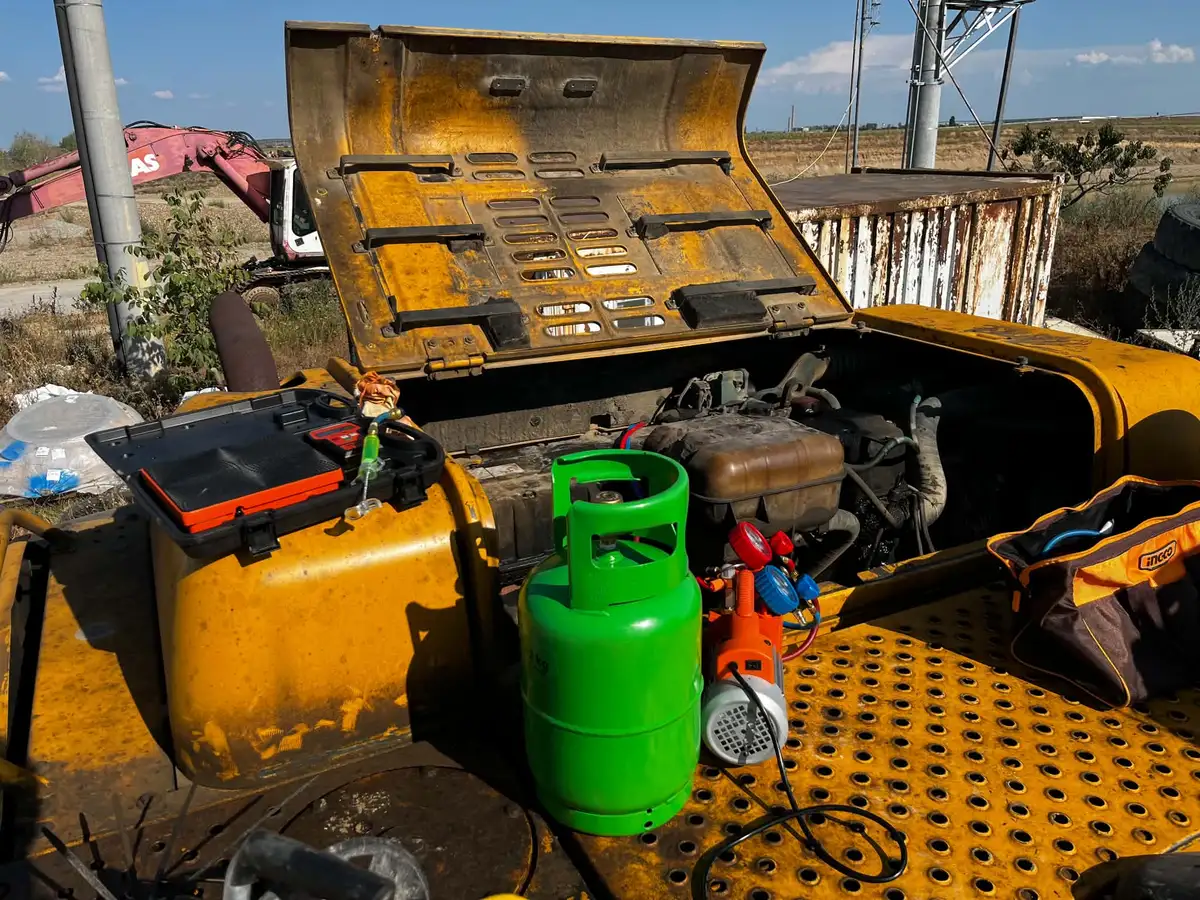 Butelie verde, balanță, pompă de vid portocalie și manometre așezate pe capota unui excavator galben, cu motorul deschis în spate
