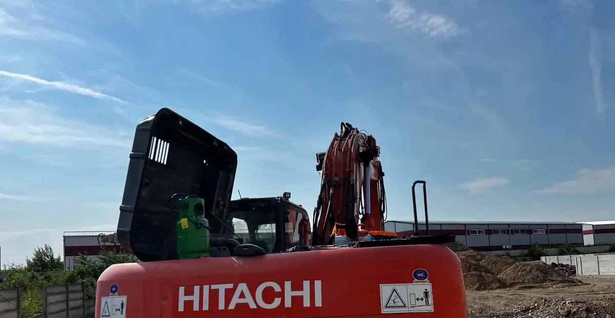 Butelie verde de agent frigorific așezată pe capota deschisă a unui excavator roșu Hitachi, sub un cer albastru