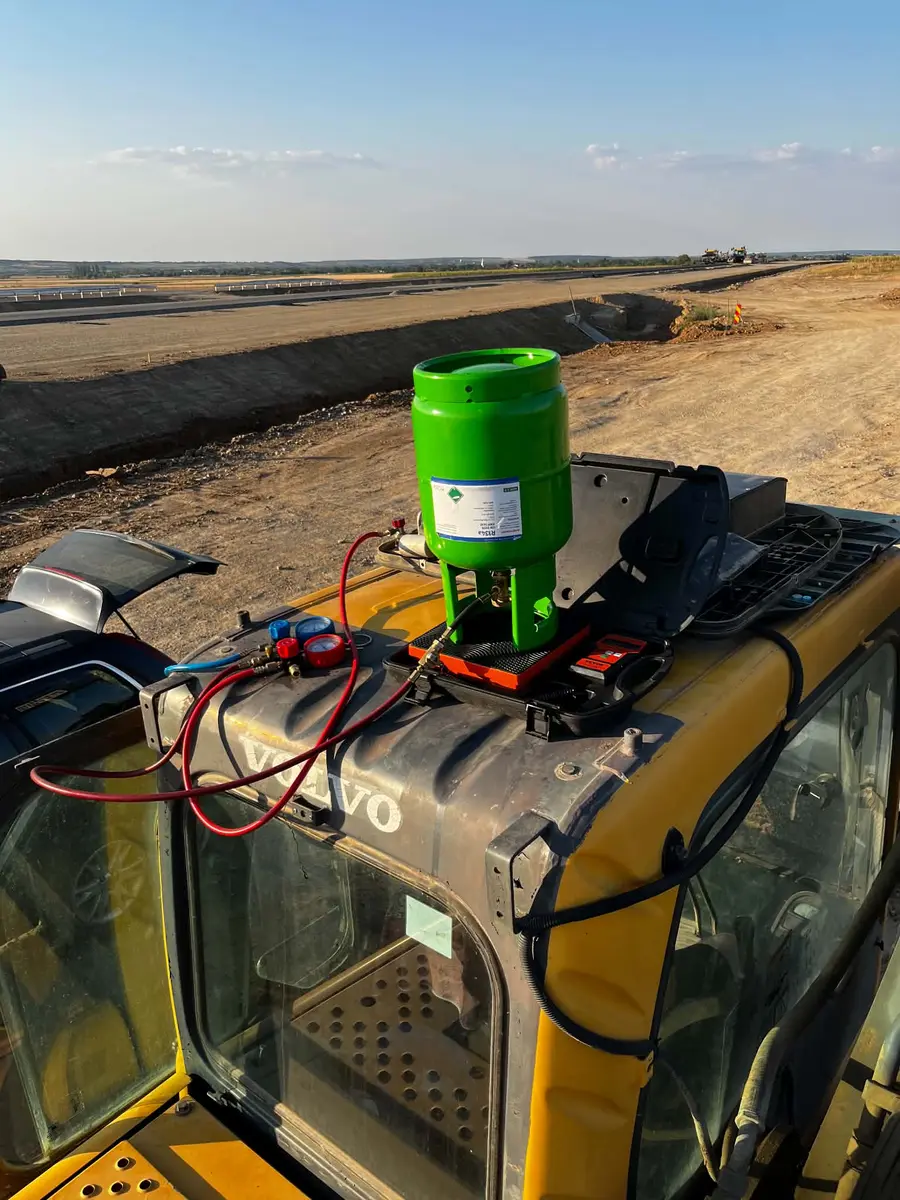 Butelie verde de R134a pe o balanță electronică, cu manometre racordate, pe cabina unui excavator Volvo, lângă un drum în lucru