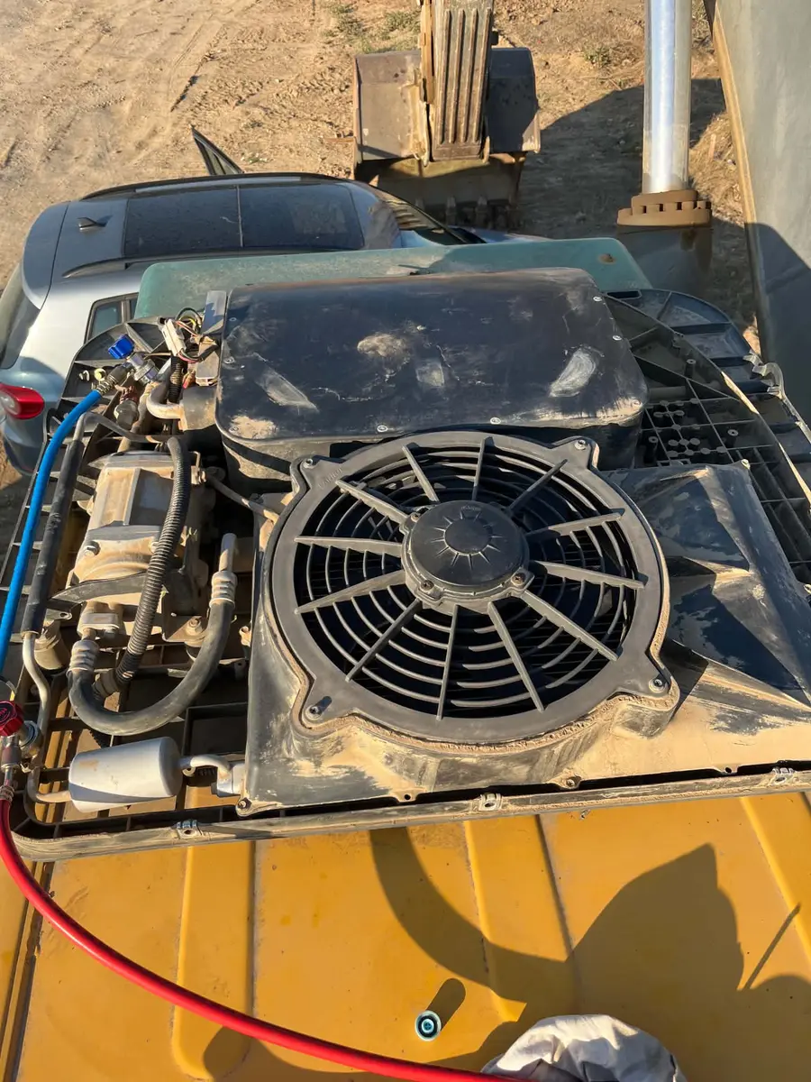 Unitatea de climatizare de pe acoperișul unui excavator galben, cu ventilatorul la vedere, compresorul și furtunurile racordate