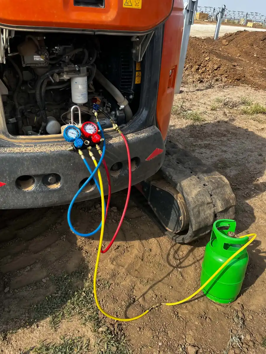 Manometre cu furtunuri roșu, albastru și galben racordate la un miniexcavator cu capota din spate deschisă, cu o butelie verde pe pământ