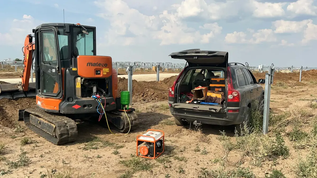 Miniexcavator Mateco portocaliu și un autoturism cu portbagajul deschis, lângă un generator portocaliu, pe un teren deschis