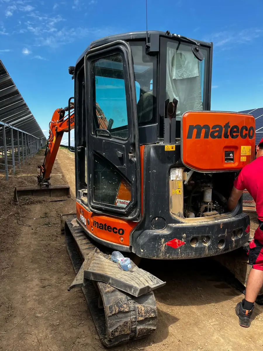 Miniexcavator Mateco văzut din spate, cu capota deschisă, pe un teren cu panouri în stânga și un om în tricou roșu în dreapta