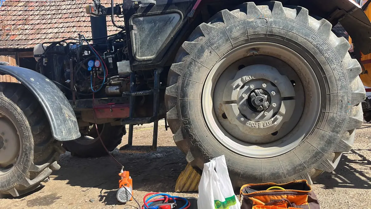 Tractor cu capota deschisă, cu manometre racordate în motor, lângă roata mare, cu o pompă de vid și furtunuri pe pământ
